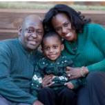 A family in matching outfits sits on the ground, smiling together with their young son in a joyful portrait